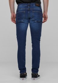 Jeans de mezclilla azul oscuro con corte slim, que cuentan con dos bolsillos traseros y una etiqueta en la cinturilla. Llevados con zapatos negros y una camiseta negra lisa.