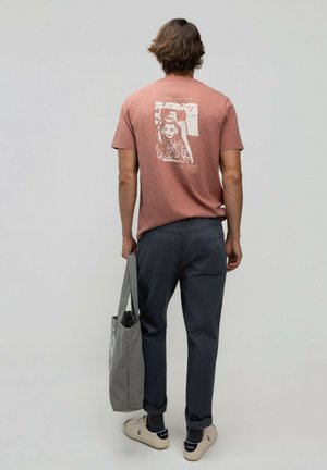 Hombre que lleva una camiseta gráfica de color óxido, pantalones oscuros y zapatillas blancas, sosteniendo una bolsa gris, de pie frente a un fondo liso.