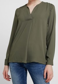 Oliven grøn bluse i let materiale med v-udskæring og frontplissé, lange ærmer og en afslappet silhuet, parret med blå jeans.
