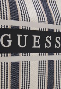 Randigt tyg i vitt, marinblått och beige. En stor svart panel med vit text som stavar "GUESS" centrerad över designen.