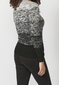 Femme portant un pull en maille dégradé noir et blanc et un jean noir, debout devant un fond clair uni, légèrement de profil.