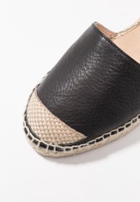 Zapato negro de cuero tipo slip-on con un acento de yute trenzado en la punta, que presenta un acabado texturizado y detalles en beige en los bordes.