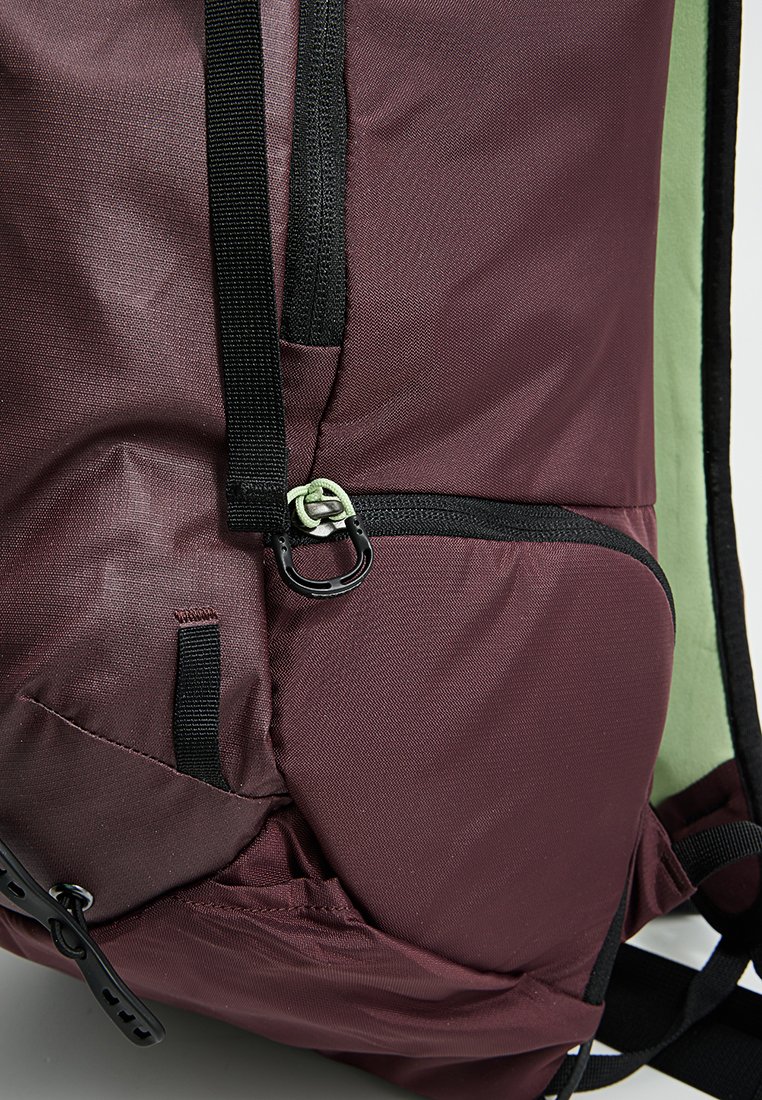 Mochila de tela color marrón con interior verde, bolsillo con cremallera, detalles negros, correas ajustables y un cierre para un asegurado seguro.