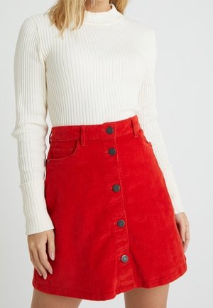 Une femme porte un pull blanc côtelé rentré dans une jupe en jean rouge vif avec des boutons sur le devant.