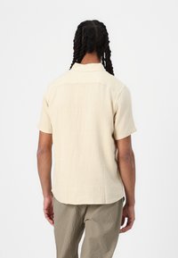 Kortærmet skjorte i lyst beige stof med en tekstureret finish. Har en klassisk krave og en lige kant, synlig rygstykke-detalje.