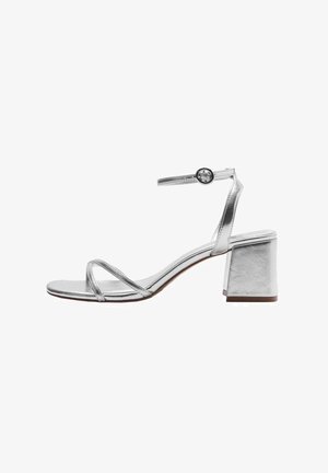 Srebrna metalna sandala z zaponko okoli gležnja in okroglo kovinsko podrobnostjo. Ima blokasto peto in tri ozke trakove na prstih.