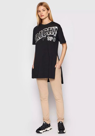 Donna che indossa una maglietta lunga nera con testo argentato, pantaloni beige e sneaker bianco e nere, in piedi con le mani in tasca.