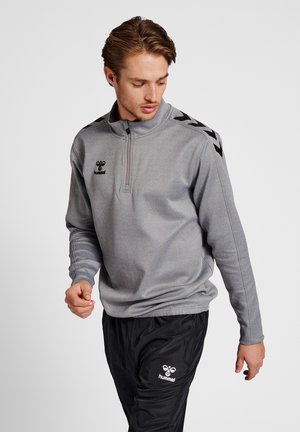 Homme en sweat-shirt gris à fermeture éclair quart de zip et pantalon de survêtement noir avec logos hummel, regardant vers le bas et tournant légèrement vers sa gauche.
