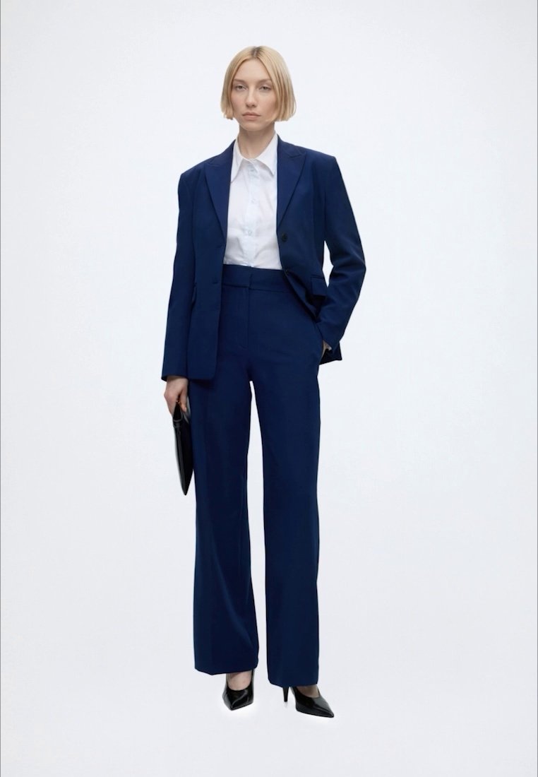 Femme debout portant un tailleur-pantalon bleu marine avec une chemise blanche et des talons noirs, tenant une pochette noire sur un fond uni.