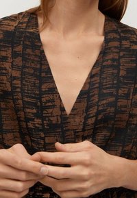 Persona con una blusa de cuello en V marrón y negro con un patrón, con las manos entrelazadas suavemente a la altura de la cintura.