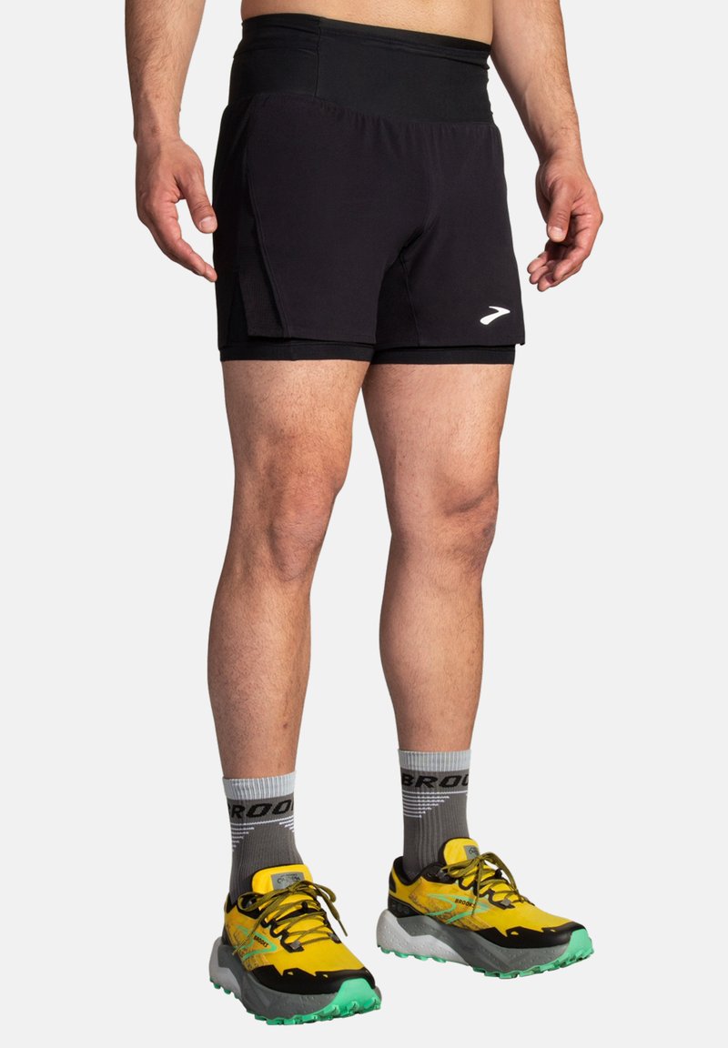 Zwarte sportshorts met een gestructureerde tailleband, zijzakken en een reflecterend logo. Gepaard met gele en zwarte hardloopschoenen en grijze sokken.