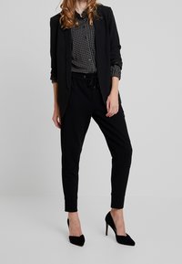 Blazer negro, camisa de lunares y pantalones jogger negros. Corte slim, tejido texturizado, cintura con cordón, combinados con tacones altos negros.