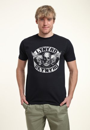 Jeune homme portant un t-shirt noir Lynyrd Skynyrd avec un motif de crâne et d’ailes, et un pantalon kaki, debout les mains dans les poches.