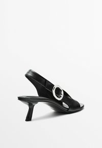 Zwarte leren slingback haksandalen hebben een uitsnijdingenontwerp, een glanzende ronde gesp als accent en een puntige neus. De hakken zijn ongeveer 3 inch.