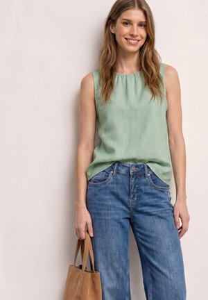 Junge Frau lächelt, trägt ärmellose hellgrüne Bluse und blaue Jeans, hält eine braune Lederhandtasche an ihrer Seite.
