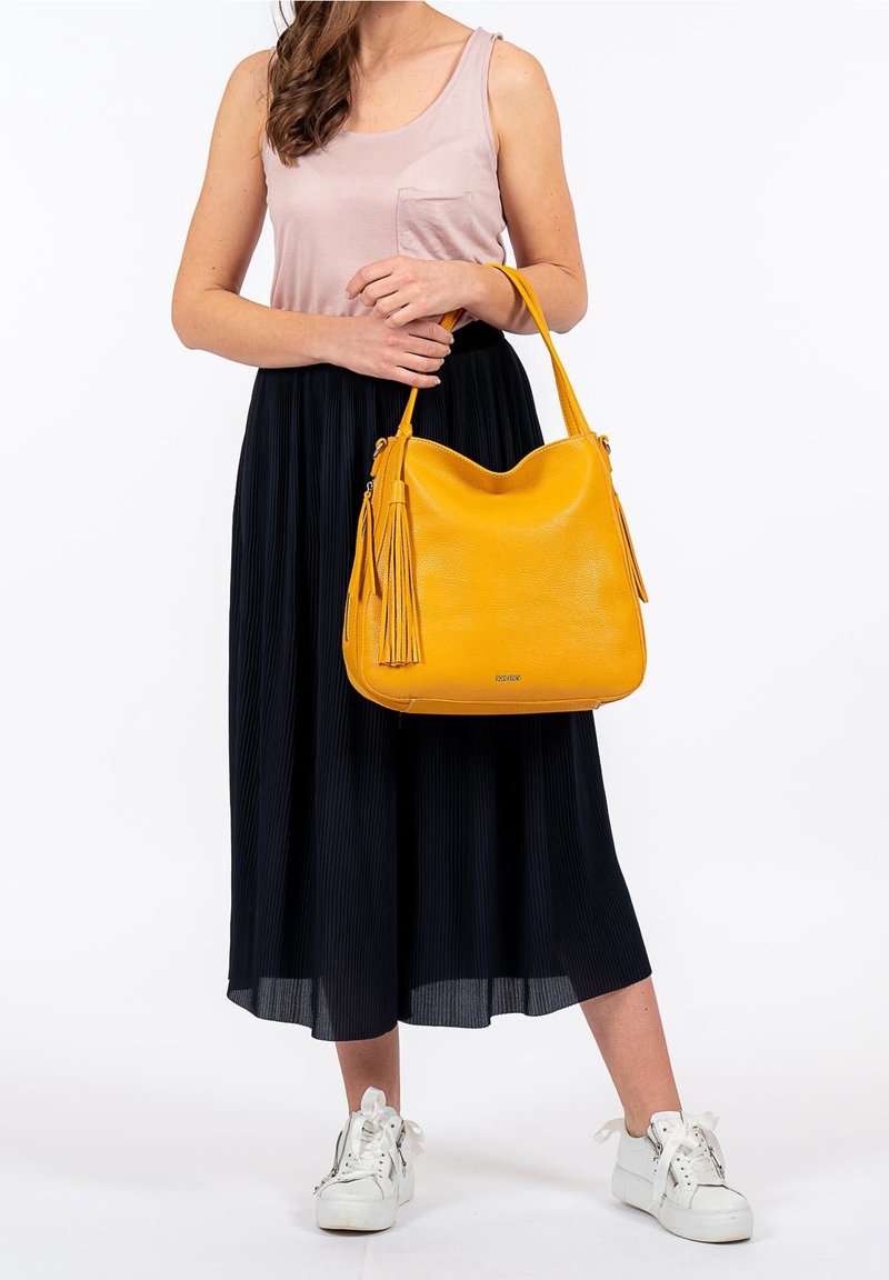 Femme en haut rose sans manches et jupe noire, tenant un grand sac à bandoulière jaune à franges, portant des baskets blanches.