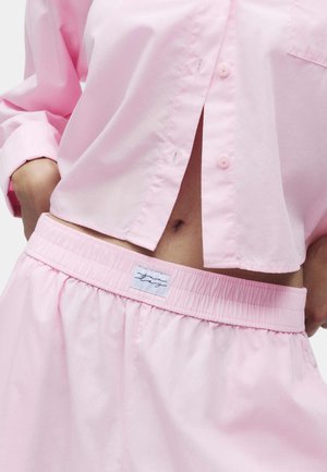 Personne portant une chemise rose pâle boutonnée et un pantalon à taille élastique assorti, montrant le ventre avec le nombril visible.