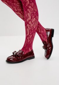 Lesklé burgundské loafers s mašlí, černou podrážkou, nošeny s růžovými krajkovými punčochami se květinovým vzorem.