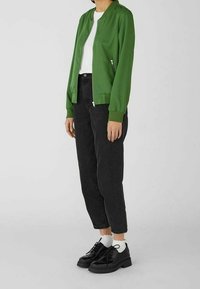 Groene bomberjack met een ritssluiting aan de voorkant, gedragen over een wit shirt. Gepaard met zwarte tapered jeans en zwarte veterschoenen.