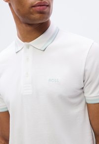 Λευκό πόλο πουκάμισο από υφασμάτινη υφή, με γιακά με ανοιχτό μπ λε ρίγες, τρία κουμπιά και κεντημένο λογότυπο "BOSS" στο στήθος.