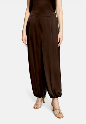 Pantalon large marron foncé avec poignets élastiqués, porté avec des sandales à talons hauts dorées à brides, sur une personne debout.