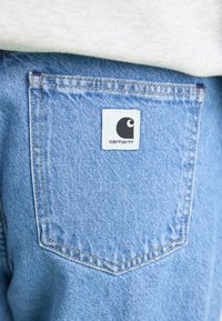 Light blue denim jeans feature a back pocket with a white label displaying the Carhartt logo. Subtle stitching details enhance the design.