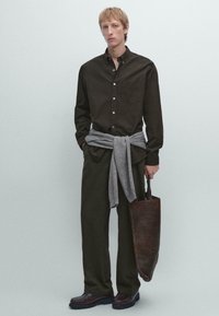 Giovane uomo che indossa una camicia e pantaloni marrone scuro con un maglione grigio legato in vita, mentre tiene una borsa tote in pelle marrone scuro.