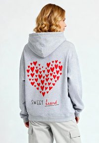 Felpa grigia con un grande grafico a forma di cuore rosso sul retro e il testo "SWEET heart". Dotata di cappuccio con cordini e vestibilità comoda.