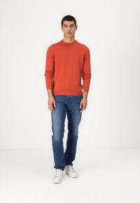 Suéter laranja tricotado, com decote redondo, punhos e bainha canelados; combinado com jeans azuis de denim e tênis brancos, posicionado contra um fundo liso.