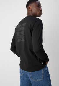 Černé tričko s dlouhým rukávem a velkým lesklým grafickým potiskem na zádech. Vyrobeno z hladké tkaniny, nabízí volný střih a kulatý výstřih.