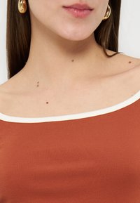 Γυναίκα που φοράει τοπ χωρίς ώμους σε σκουριά απόχρωση με λευκή λεπτομέρεια, χρυσούς κρίκους στα σκουλαρίκια και εμφανή σπίλους στον λαιμό και στους ώμους.