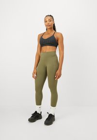 Fit kvinna med flätad frisyr står iförd en svart sportbehå, olivgröna högmidjade leggings med ficka, vita Nike-strumpor och svarta sneakers.