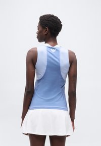 Γυναίκα με κοντά μαλλιά, φορώντας αθλητικό μπλουζάκι χωρίς μανίκια μπλε χρώματος και λευκή φούστα, εμφανίζεται από πίσω μπροστά σε απλό φωτεινό φόντο.