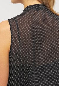 Mujer que lleva una blusa sin mangas y transparente de color negro con pequeños lunares blancos, con el escote y la parte superior de la espalda visibles, cabello castaño claro.