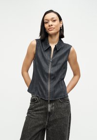 Αμάνικο μπλε τζιν μπουφάν με φερμουάρ μπροστά, μυτερό γιακά και πλαϊνές ραφές, συνδυασμένο με ψηλόμεσο σκούρο τζιν παντελόνι. Λείο ύφασμα.