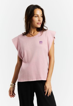 Žena v růžovém tílku bez rukávů s malým fialovým logem na hrudi, oblečená v černých kalhotách a zlatých náramcích, dívající se stranou.