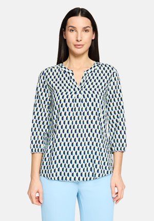 Langärmelige Bluse mit einem geometrischen Muster in Marineblau und Hellblau auf weißem Hintergrund. V-Ausschnitt und gesammelte Bündchen.