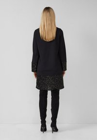 Kobieta z długimi blond włosami, ubrana w czarny płaszcz z cekinowymi mankietami i dołem, w połączeniu z czarnymi wysokimi kozaczkami do kolan, stojąca.