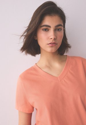 Nuori nainen, jolla on lyhyet ruskeat hiukset ja punertavan korallinvärinen V-kaula-aukkoinen T-paita, seisoo vaaleanpunaisen taustan edessä.