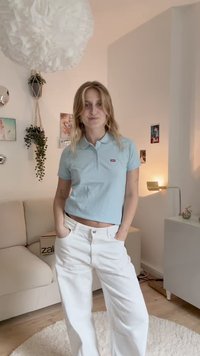 Vaaleansininen polo-paita, jossa on kaulus ja pieni punainen logo, yhdistettynä valkoisiin leveisiin housuihin. Huoneessa on moderni sisustus.