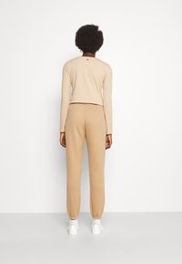 Beżowa bluzka z długim rękawem i pasującymi spodniami z elastycznymi mankietami. Miękka tkanina, krótki fason, minimalistyczne detale i subtelne logo z tyłu.