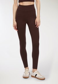 Leggings marroni realizzati in tessuto elasticizzato con un alto punto vita, abbinati a sneakers bianche con dettagli in marrone e nero.