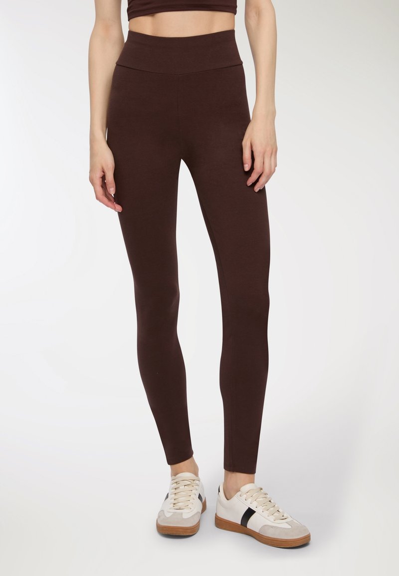 Leggings marroni realizzati in tessuto elasticizzato con un alto punto vita, abbinati a sneakers bianche con dettagli in marrone e nero.