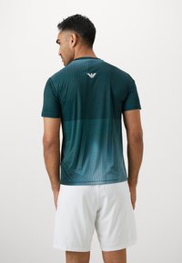 Sötét teal színű sportpóló, amelynek színátmenetes mintáján pontok és vonalak találhatóak, rövid ujjú, és a hátrész felső részén egy logóval van ellátva.