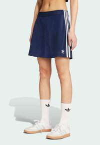 Mornarska atletska suknja s bijelim trostrukim dizajnom, lagani materijal, elastična stražnja traka i Adidasov logo na lijevom rubu.