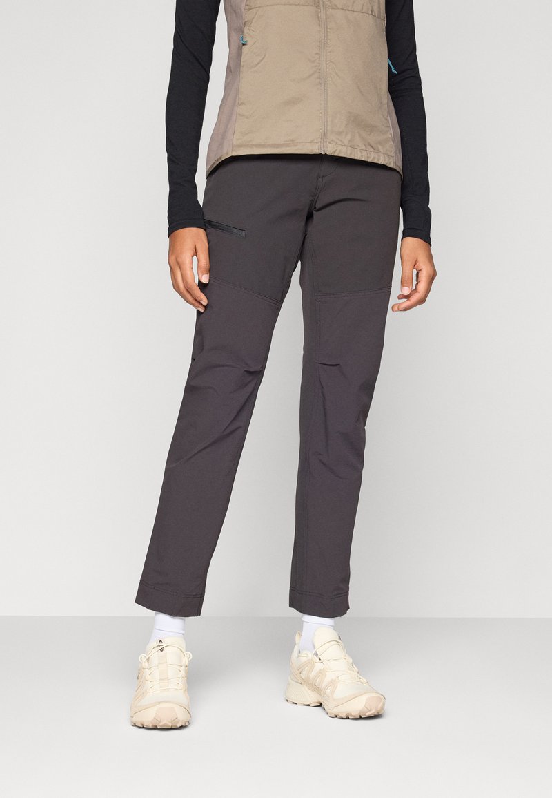 Donkergrijze outdoorbroek met gebogen knieën en zakken, gecombineerd met een beige vest, witte sokken en lichtgekleurde sneakers.