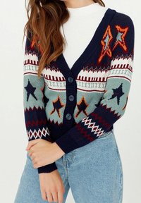 Mehrfarbiger Strickcardigan mit navyblauem Grund, hellblauen, roten und weißen Mustern, einschließlich Sterne und Zickzack-Motiven; Knopfverschluss.