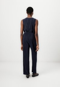 Combinaison bleu marine avec des rayures verticales, sans manches, taille élastique et pantalons larges. Des ballerines noires complètent la tenue.