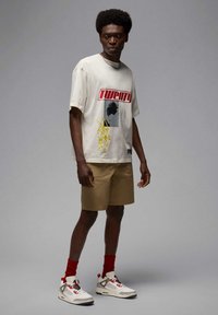 Creme t-shirt med grafisk tryk og gule accenter, parret med lysebrune shorts. Sneakers har grå, røde og sorte detaljer. Røde sokker er synlige.