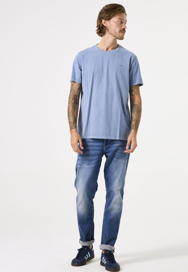 Camiseta azul de cuello redondo en algodón suave, combinada con jeans azul claro y zapatillas de deporte azul marino. Se ven patrones de tatuajes detallados en los brazos.